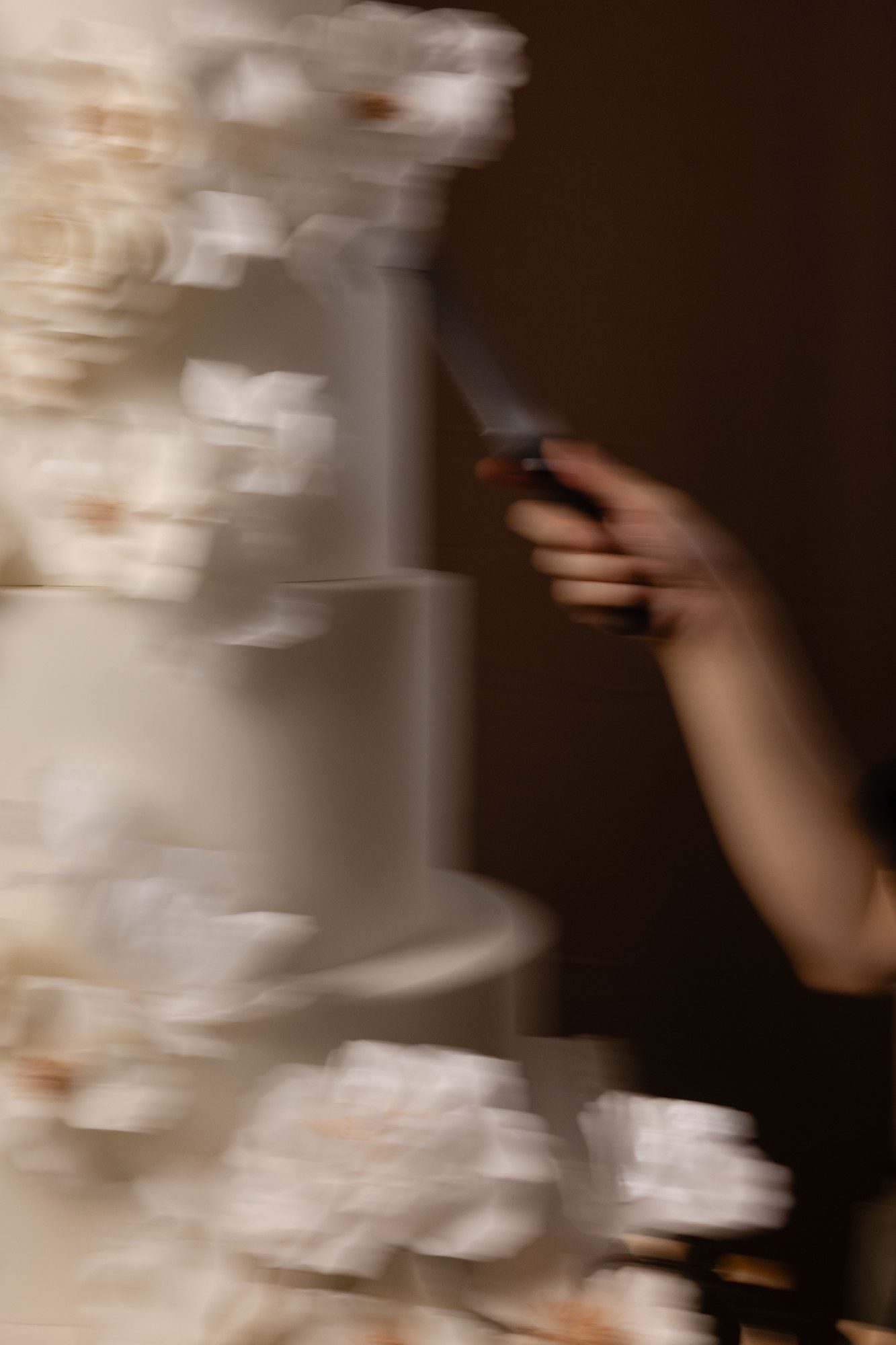 A hand with a cake knife beside a tall floral wedding cake, in motion.