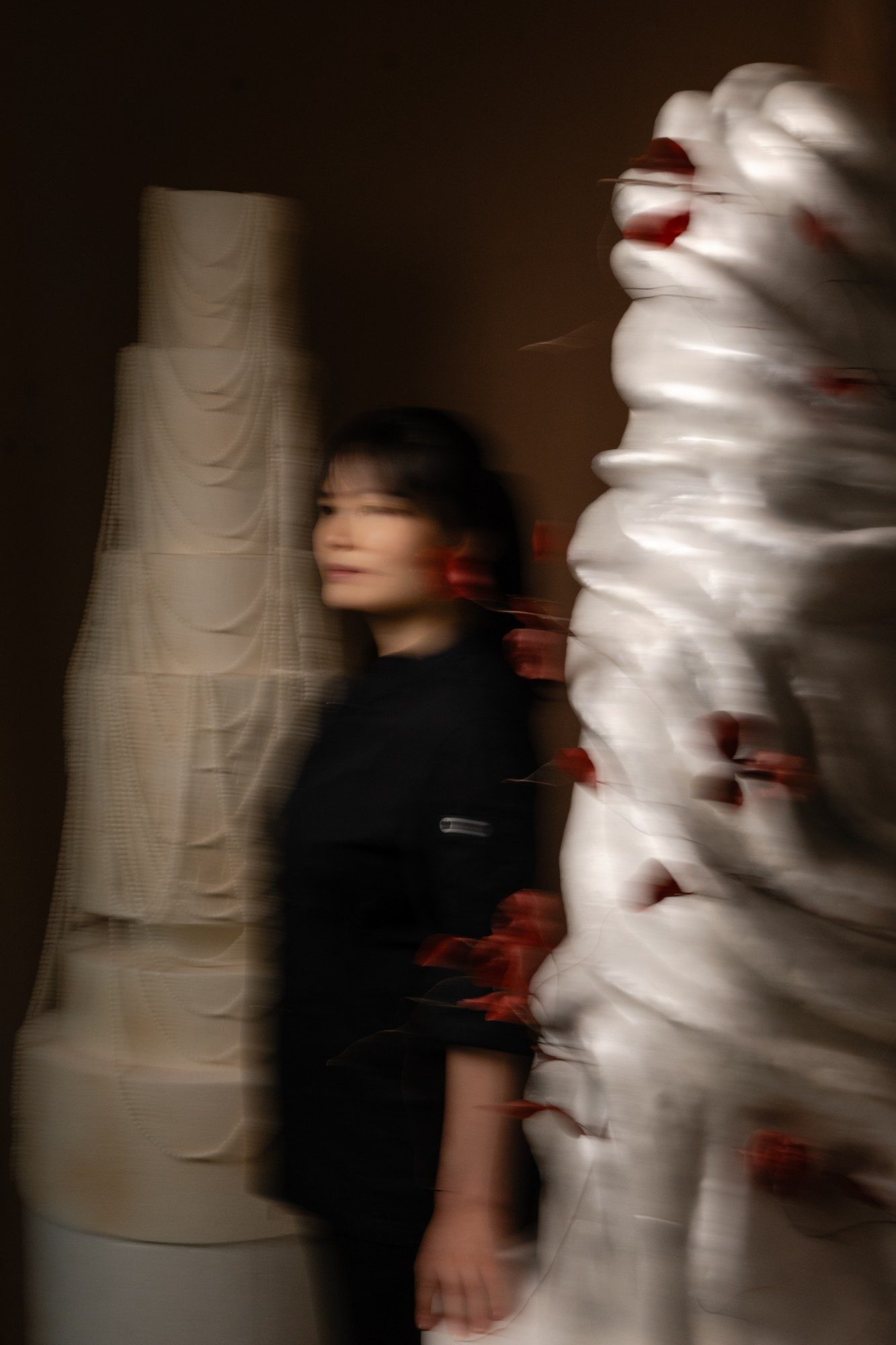 Fiona Lewy, blurred in motion, between a pearl-draped wedding cake and a silver sculptural cake with oxblood petals.