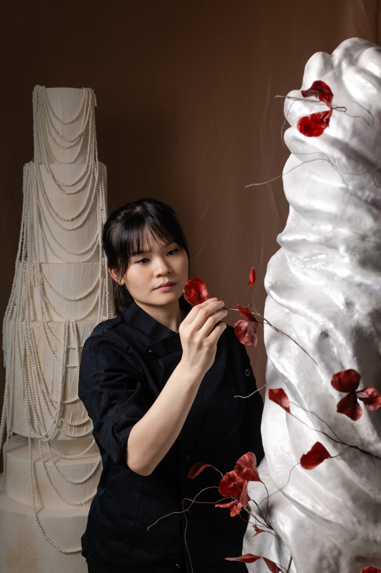 Fiona Lewy placing oxblood sugar petals on a silver sculptural wedding cake, a pearl-draped cake behind her.