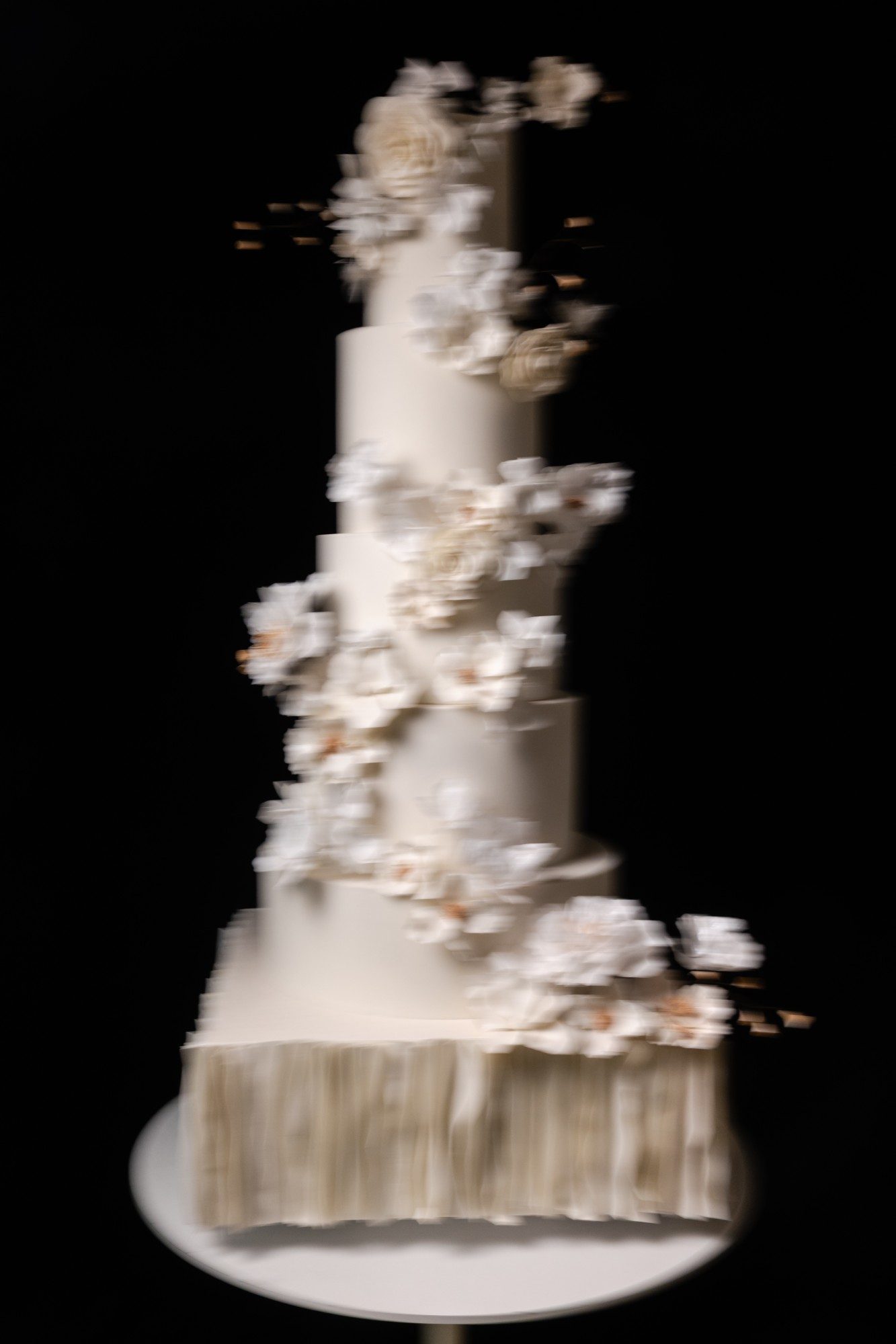 A tall ivory wedding cake with cascading sugar flowers and a pleated base, photographed in motion against black.