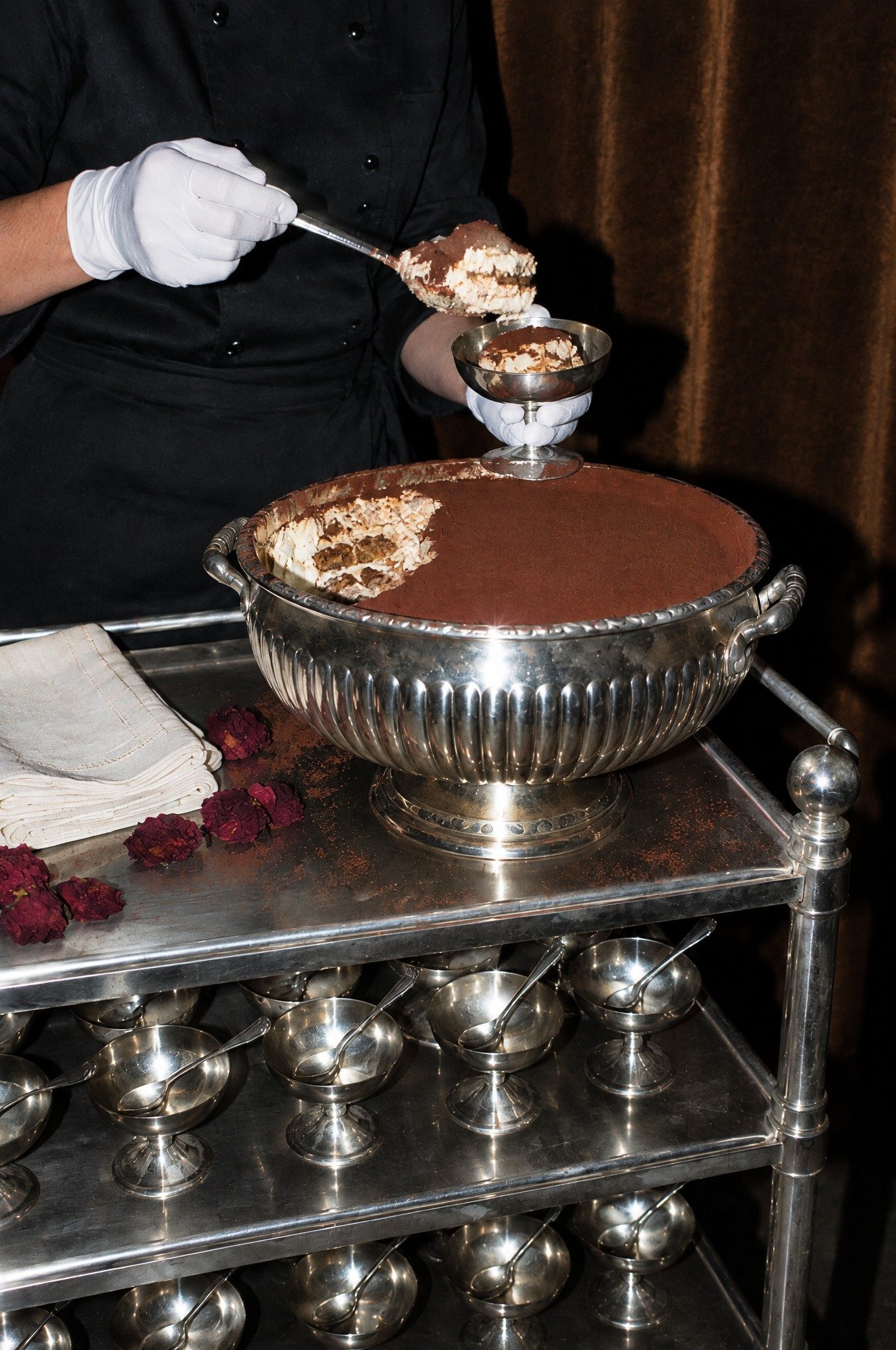 A server in white gloves scooping tiramisu from a silver tureen on a silver trolley.